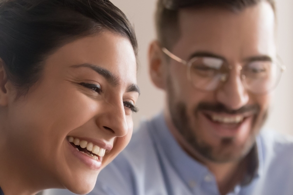 An Asian woman with short dark hair wearing a polka dot shirt laughs with a white male colleague who is wearing glasses, had a beard and has a blue shirt